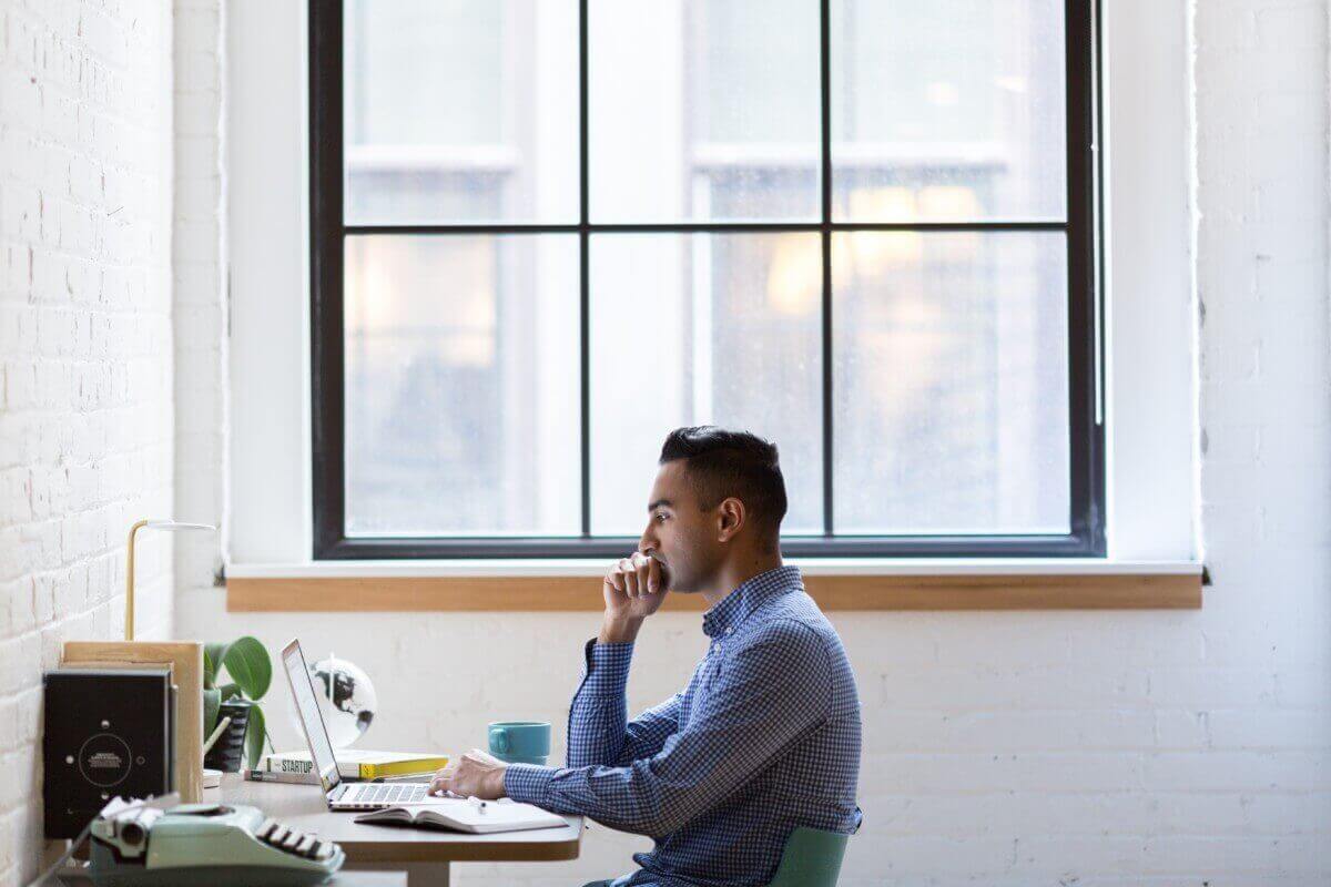 man sitting at a desk looking at a computer with a big window behind him
