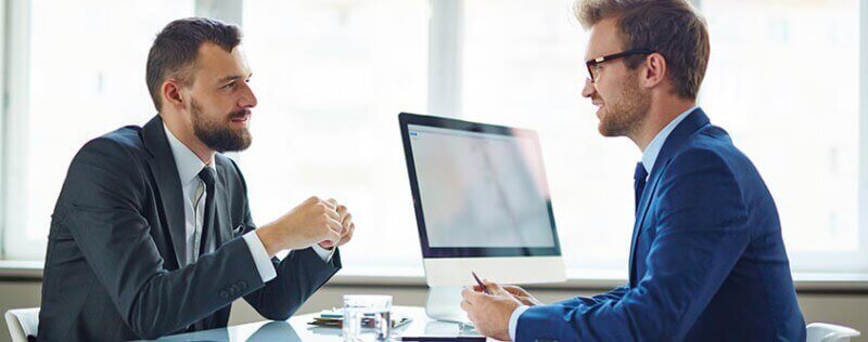 Two business professionals sitting and talking at a desk with a computer in the background
