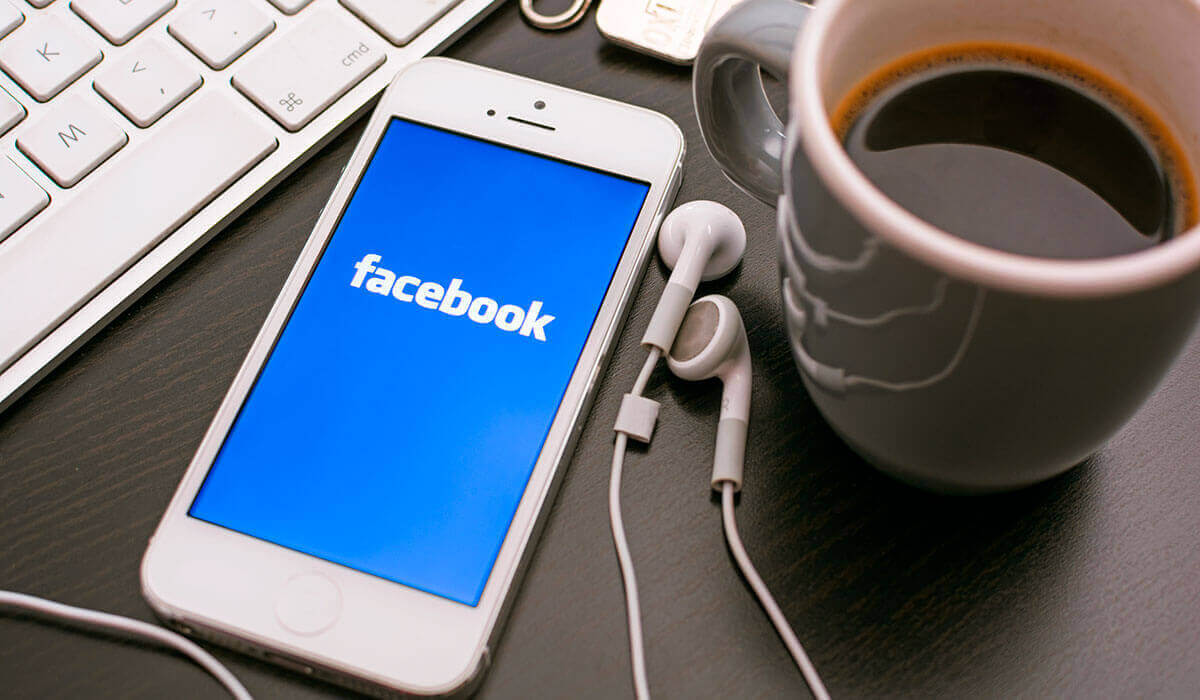 A white iPhone showing the facebook logo near a laptop, headphones, and a coffee cup on a desk