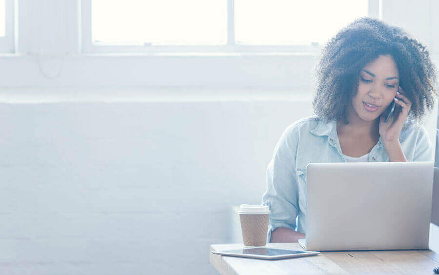 Person talking on a cell phone sitting at a desk with a laptop, to-go cup of coffee, and tablet