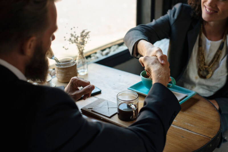 Two people sitting down and shaking hands at a cafe table with coffee drinks near a window