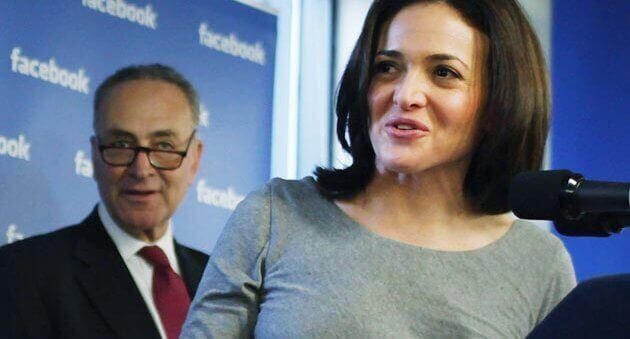 Speaker on a podium with a microphone looking to the side in front of a man and Facebook wall