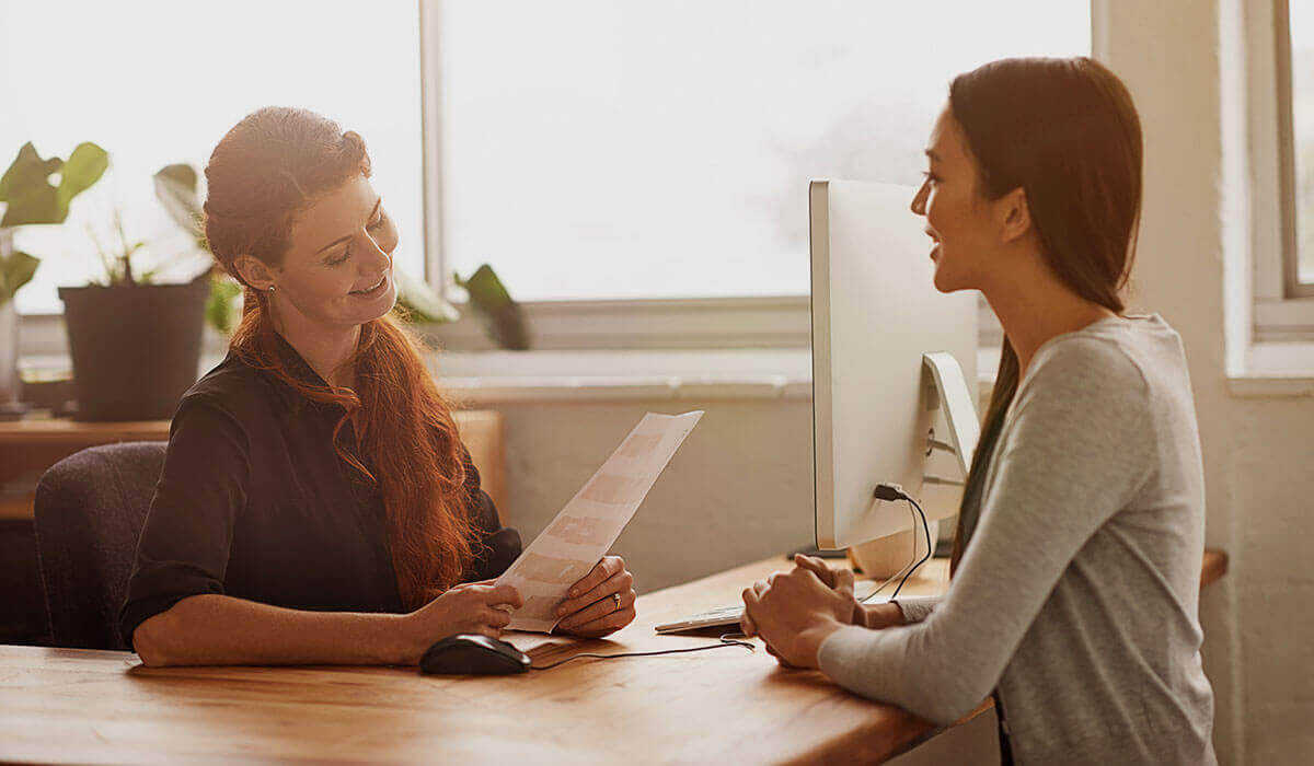 An interviewer sitting at a desk and holding a paper in front of an interviewee