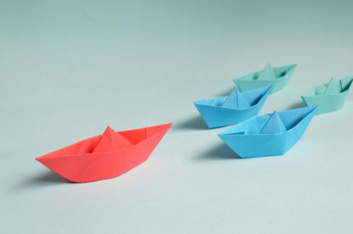 Paper boats on a table where the red one takes the lead as the leader. The image demonstrates that you can lead through collaboration.