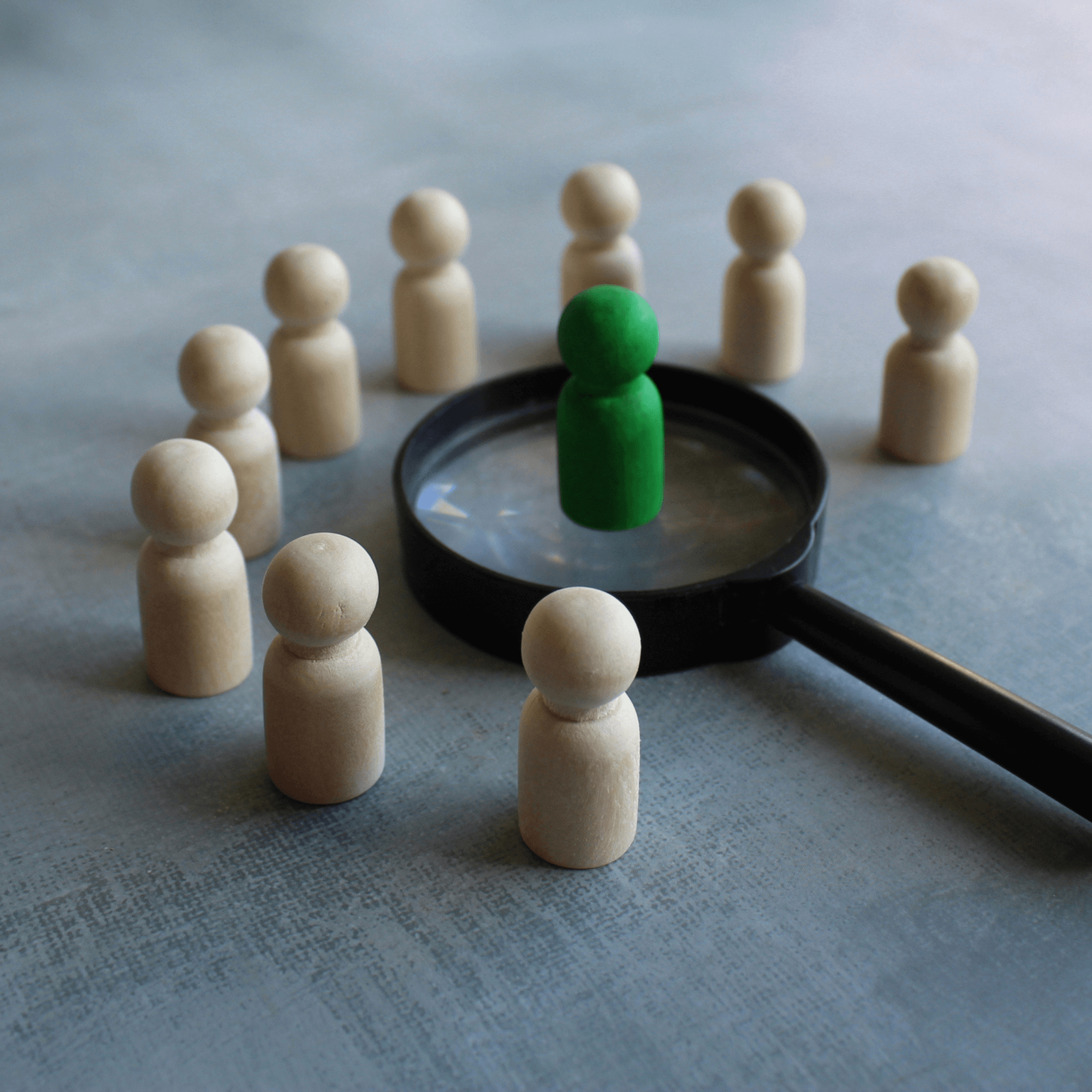 Wooden people figures standing around a magnifying glass with one standing on it.